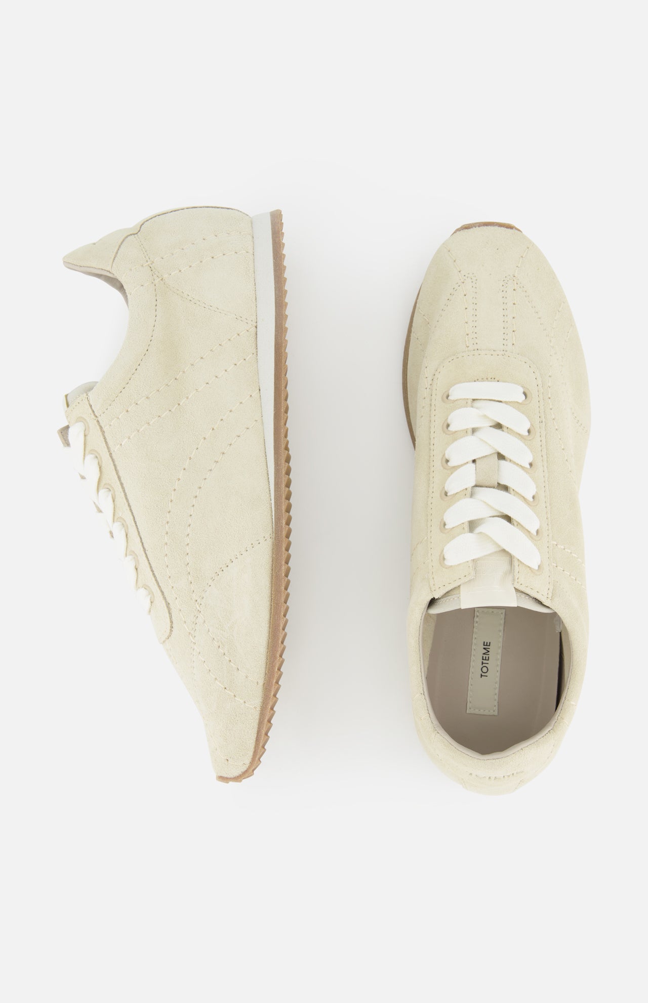 TOTEME’s Flex Suede Low Profile Sneakers, expertly crafted in Italy, are shown on a white background with white laces and brown rubber soles—one displayed side-on, the other from above.