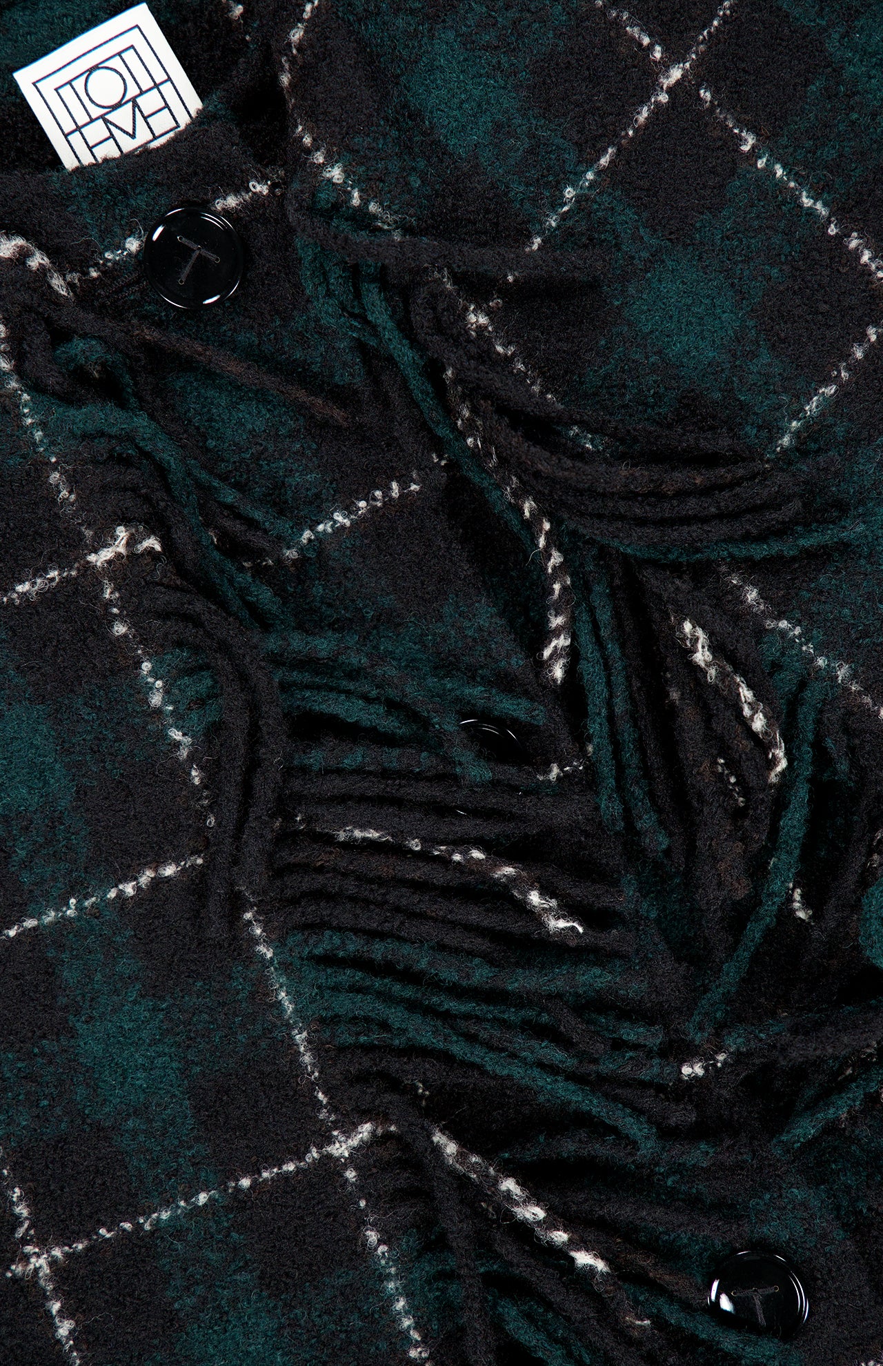 Close-up of the TOTEME Fringed Scarf Jacket, featuring a dark green and black plaid wool-blend fabric with white lines, black buttons, and fringed detailing. A white label with a geometric logo appears in the top left corner.