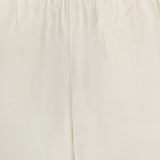 Close-up of the upper section of TOTEME Monogram Silk PJ Bottoms in cream, featuring straight legs, an elastic waistband with subtle stitched patterns, a small fabric label, and an embroidered monogram at the center.