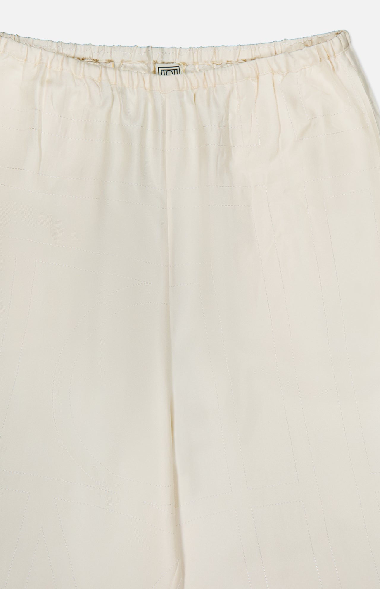 Close-up of the upper section of TOTEME Monogram Silk PJ Bottoms in cream, featuring straight legs, an elastic waistband with subtle stitched patterns, a small fabric label, and an embroidered monogram at the center.