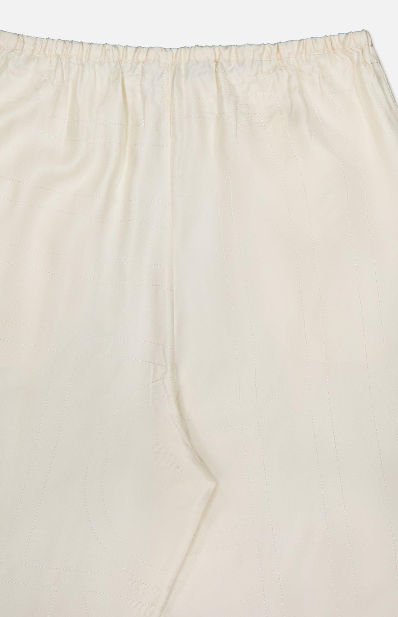 Close-up of TOTEME's Monogram Silk PJ Bottoms: cream-colored straight-leg pants featuring an elastic waistband, subtle stitching, and a delicate embroidered monogram on smooth silk fabric.