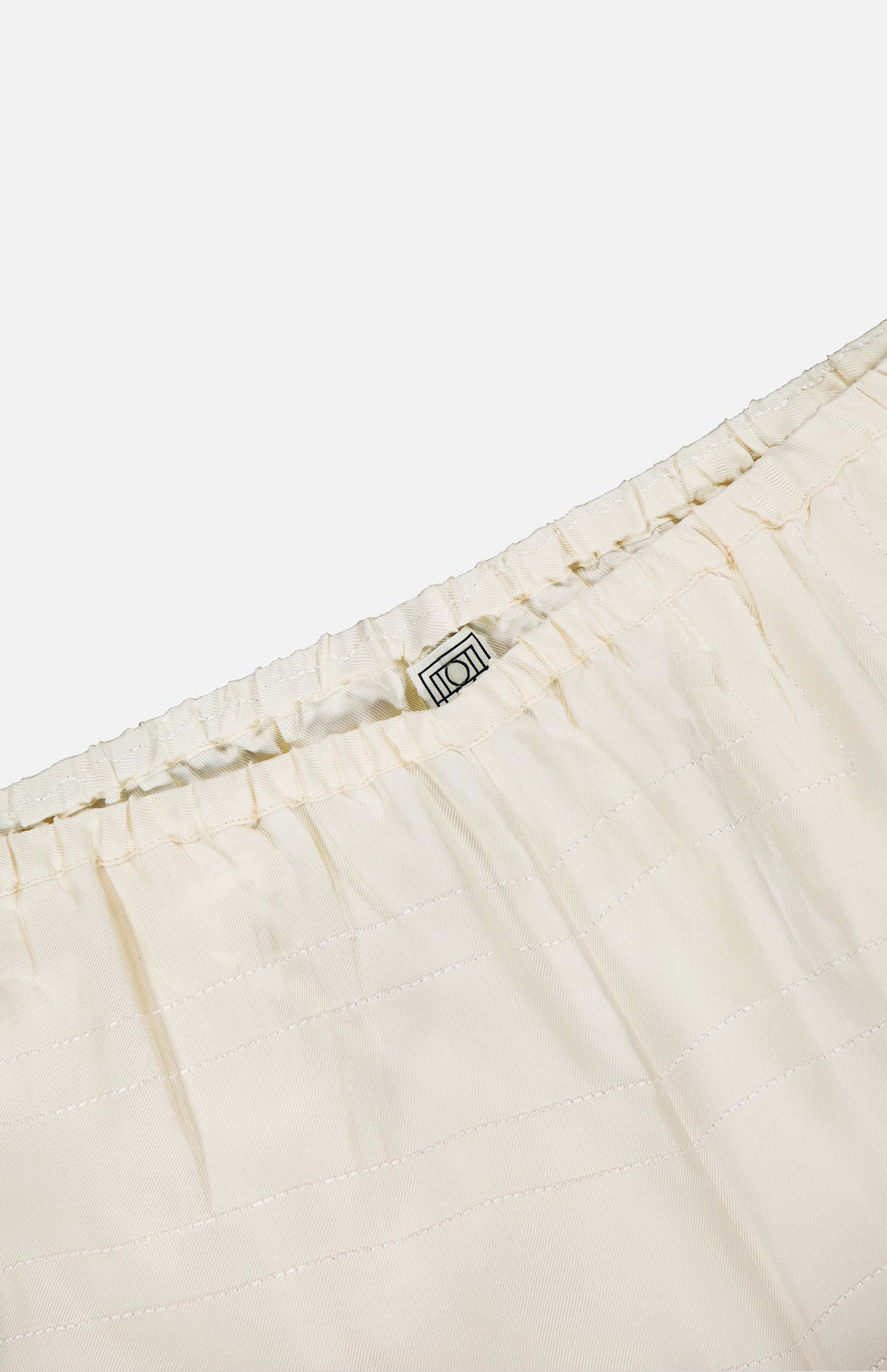 Close-up of the waistband on TOTEME's Monogram Silk PJ Bottoms, featuring cream-colored pleated silk, an elastic band, and a small fabric label sewn inside.