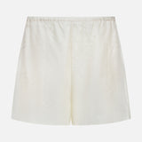 TOTEME Monogram Silk PJ Shorts feature cream satin fabric, an elastic waistband, and subtle stitched detailing. Photographed against a plain white background, these shorts evoke classic silk sleepwear styles.