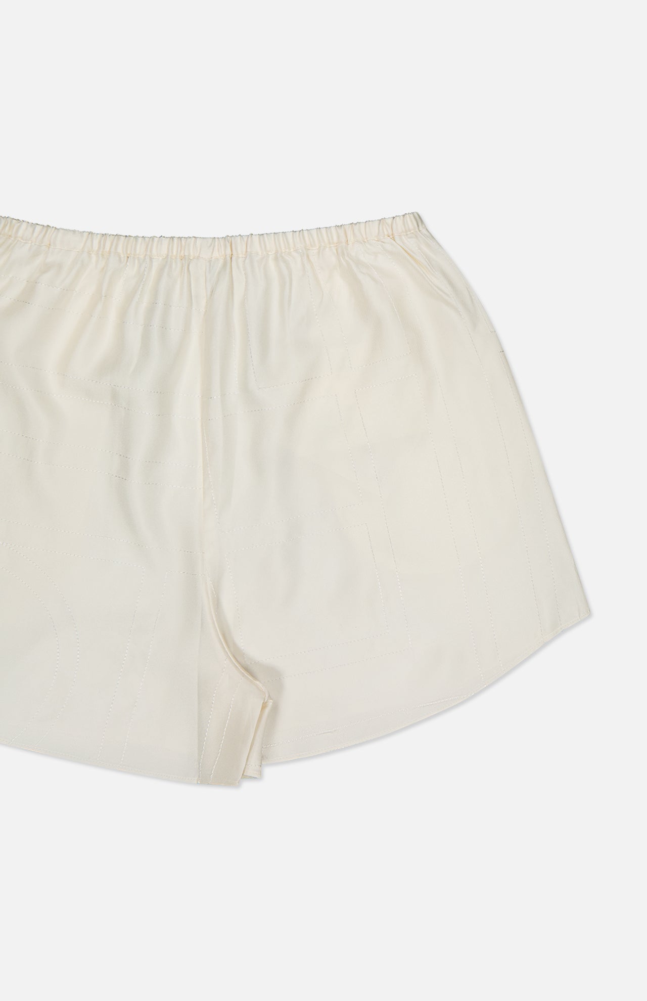A close-up of TOTEME's Monogram Silk PJ Shorts in cream, made from lightweight silk with an elastic waistband and subtle embroidered monogram, shown on a white background.