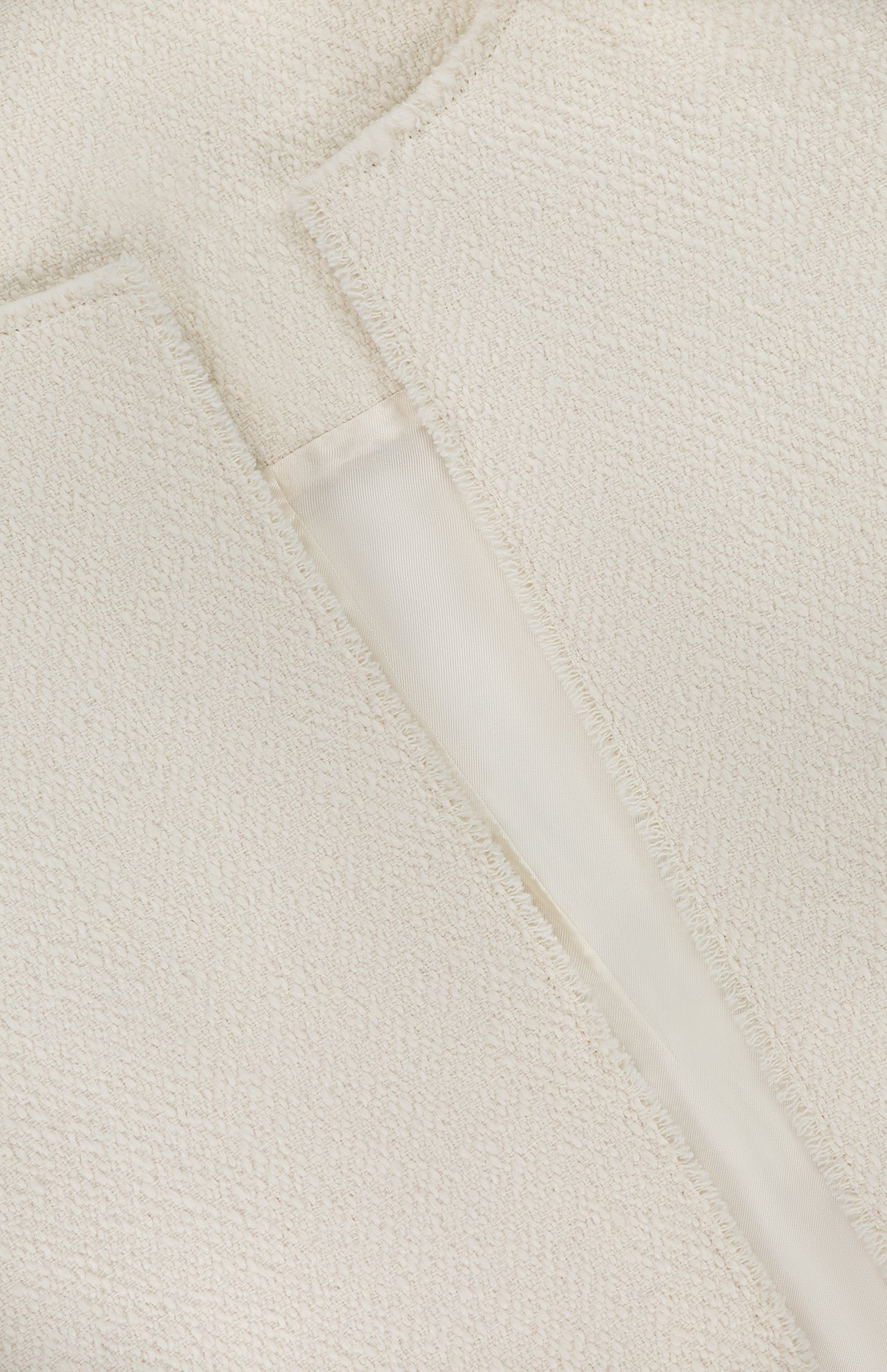 Close-up of TOTEME's Open-Front Bouclé Blazer in off-white, showing textured fabric with a visible seam and raw edge. A partially open slit reveals the smooth, lighter inner lining.