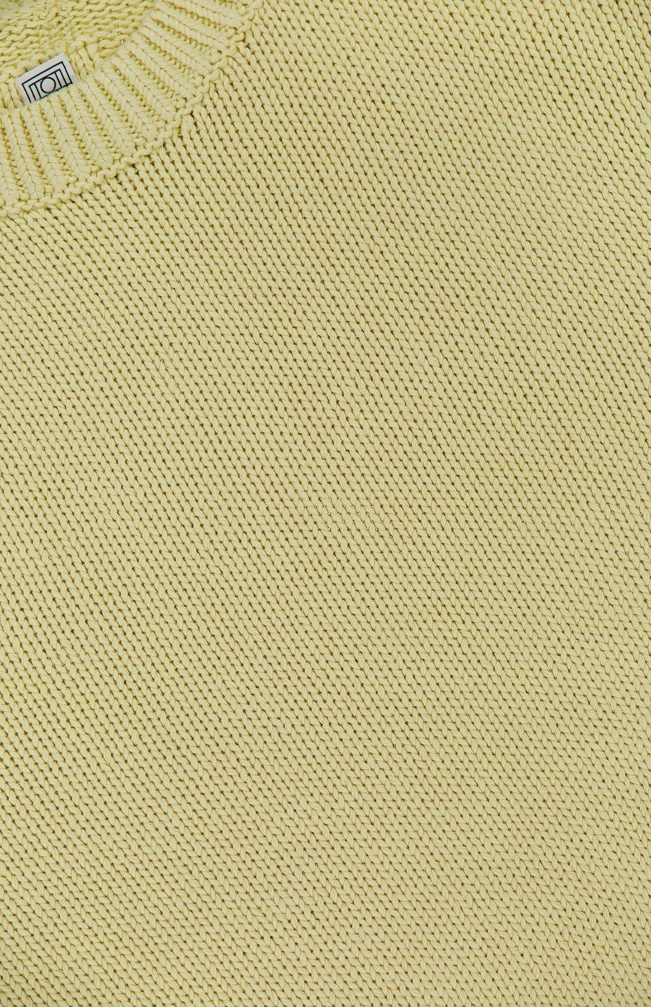 Close-up of the Oversized Crewneck Knit Sweater by TOTEME in butter yellow, showcasing textured stitching and a glimpse of the collar with a white tag.