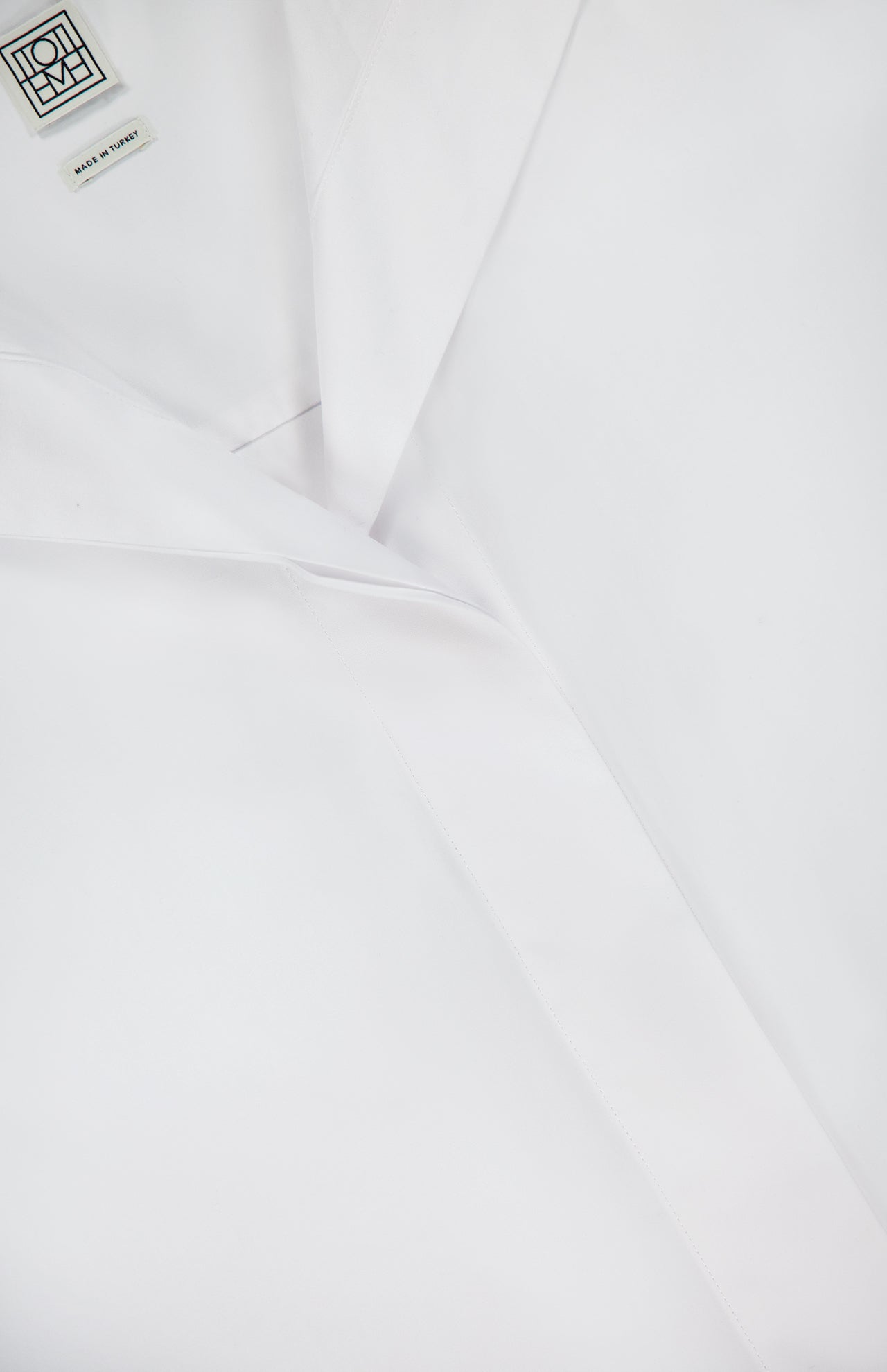 Close-up of TOTEME’s Sharp Poplin Dress Shirt in crisp white cotton, featuring a visible collar, MADE IN ITALY tag, and a concealed button-down front for subtle elegance.