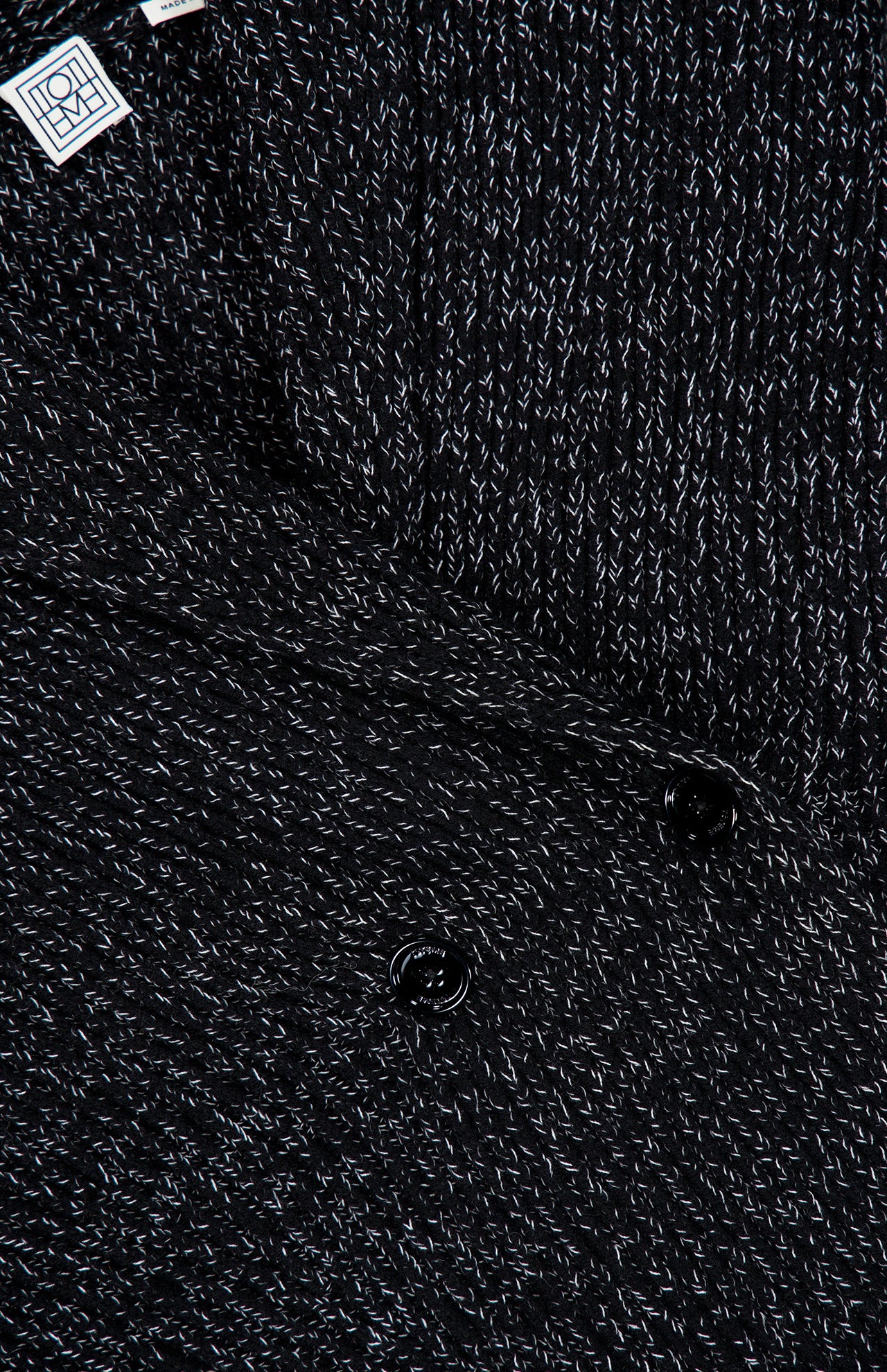 Close-up of the TOTEME Shawl-Lapel Cardigan’s black and white textured wool-blend fabric, featuring a subtle woven pattern, two black buttons along the front closure, and a small white TOTEME label in the top left corner.