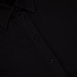 Close-up of the TOTEME Slouch Jersey Shirt Dress, featuring a black button-up front with four buttons, a collar, and a small white tag by the neckline. The featherlight jersey fabric creates a smooth, classic silhouette.
