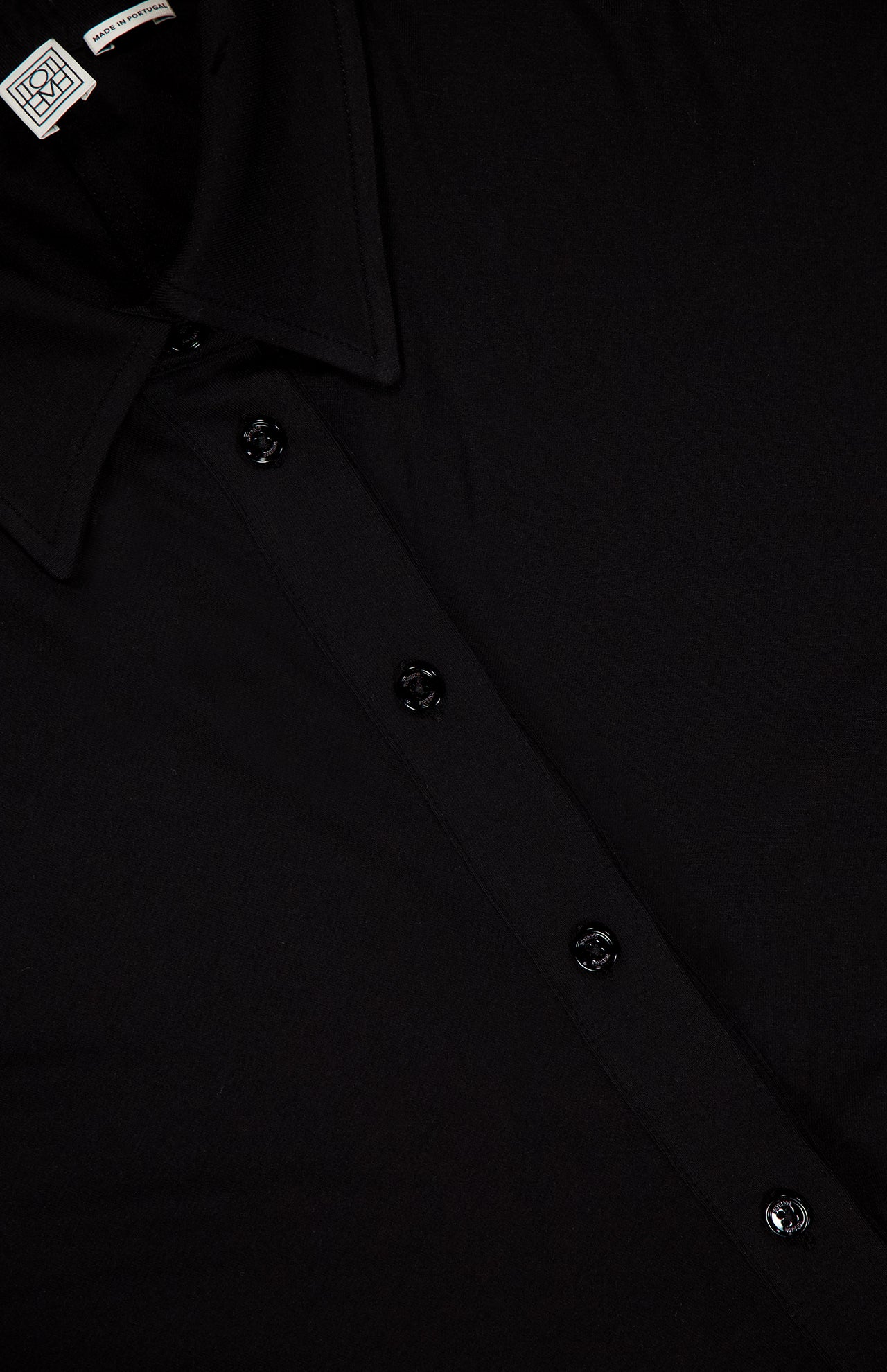 Close-up of the TOTEME Slouch Jersey Shirt Dress, featuring a black button-up front with four buttons, a collar, and a small white tag by the neckline. The featherlight jersey fabric creates a smooth, classic silhouette.