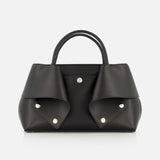 The TOTEME Small Mercer Leather Tote Bag features black leather with two short handles, silver button details, and folded flap accents on each side. Expertly crafted in Italy, this sophisticated designer tote is displayed against a plain white background.