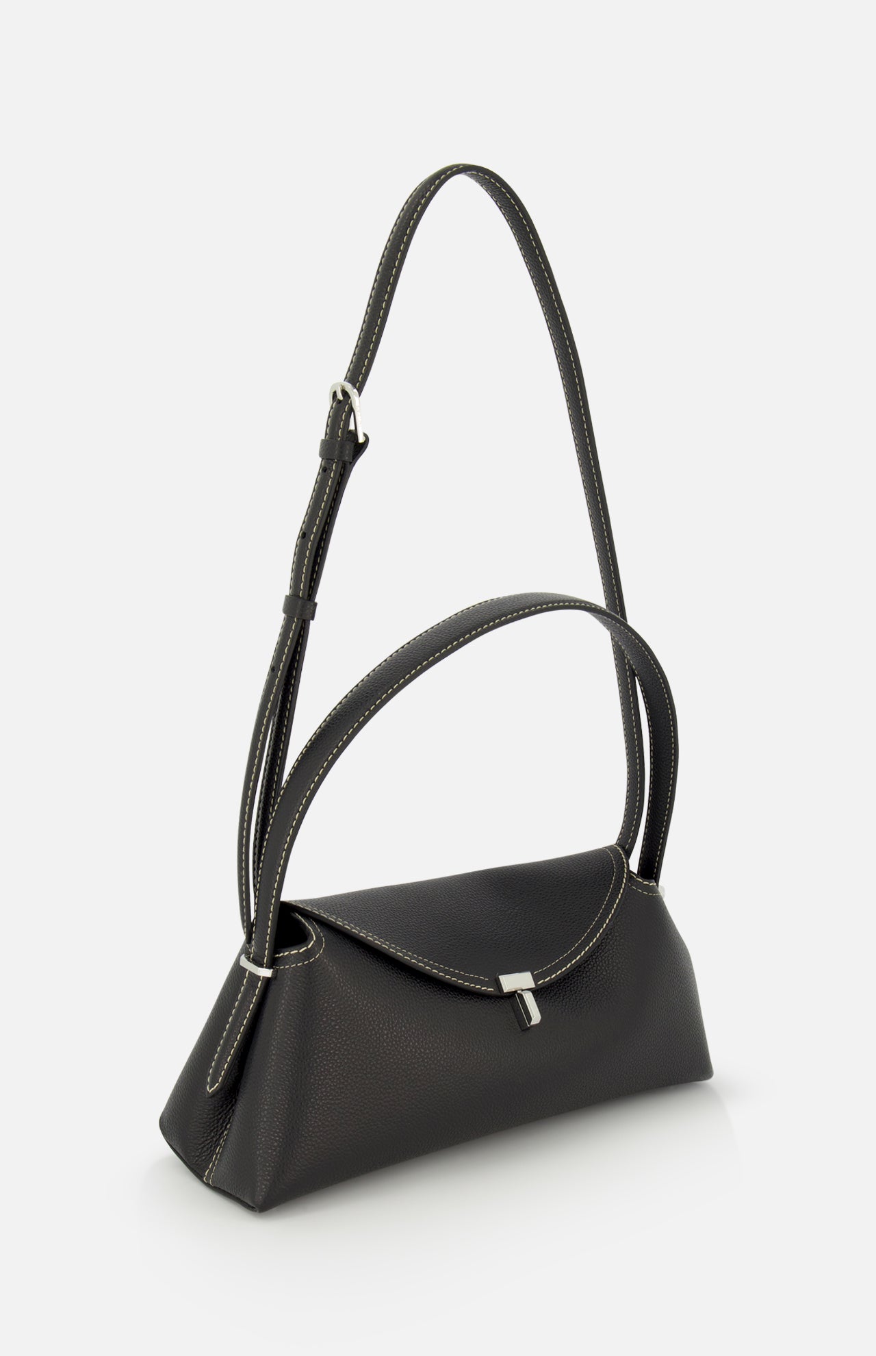 The TOTEME T-Lock Grained Leather Shoulder Bag features black pebble-grained leather, white stitching, a flap closure with a silver buckle, and an adjustable strap. Photographed on a plain white background. Made in Italy.