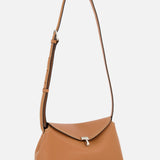 The TOTEME T-Lock Grained-Leather Clutch is a tan sculptural bag with a curved rectangular silhouette, adjustable strap, silver clasp, and white stitching, crafted in Italy and shown against a plain white background.