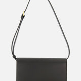 The TOTEME T-Lock Leather Crossbody Bag is a sculptural shoulder bag crafted from black leather, featuring a rectangular shape, slim profile, and an adjustable strap, beautifully displayed against a plain white background.