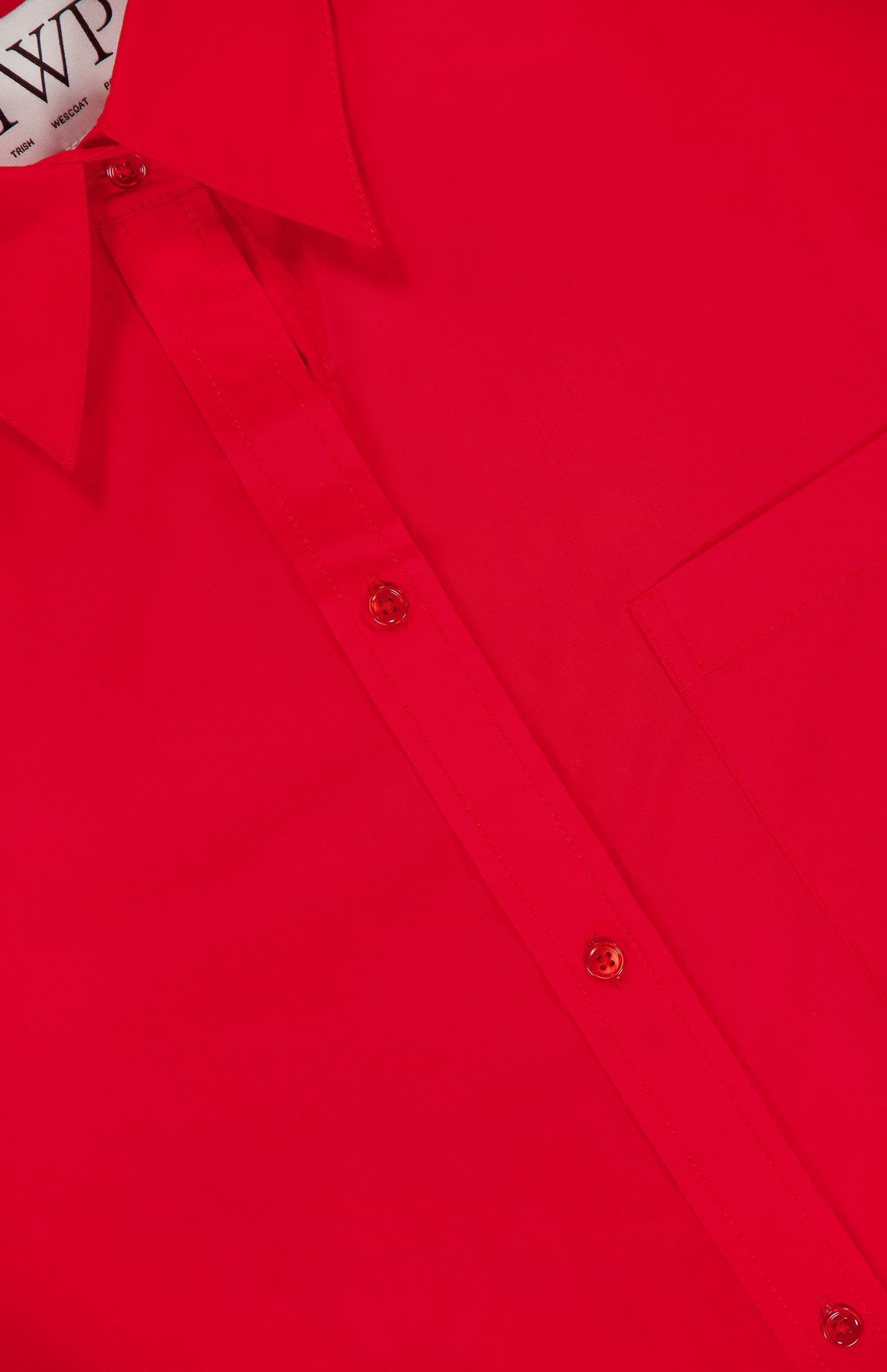Close-up of the Big Joe Oversized Shirt Dress by TWP, featuring a bright red color, button-up front, collared neckline, chest pocket, and a white tag with "WP"—the perfect comfortable workwear option for women.