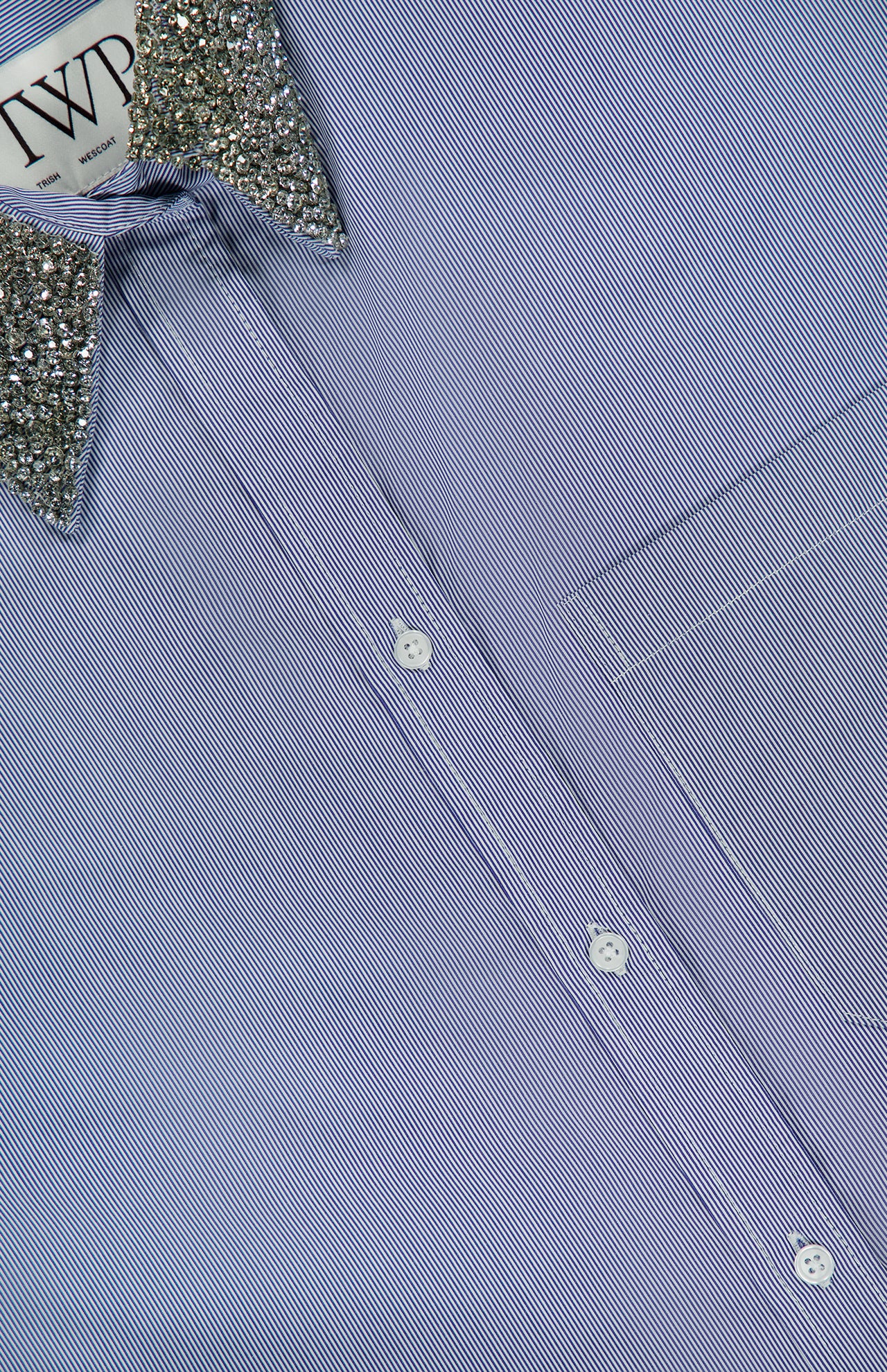 A close-up of the TWP Big Joe with Crystal Collar Shirt, an oversized blue pinstripe design detailed with a silver sequin-embellished crystal collar and a visible chest pocket—an elegant update to modern women’s workwear.