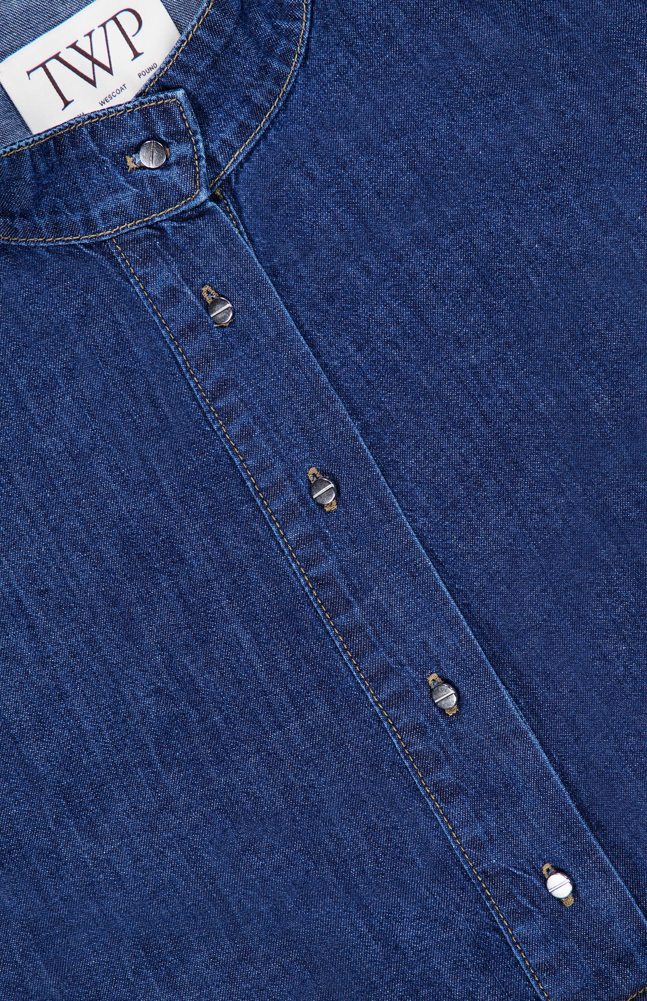 Close-up of the TWP Denim Tamzin Wrap Shirt in blue denim, showcasing a round collar, signature white TWP label, and silver snap buttons down the front—an effortless twist on classic women’s workwear.