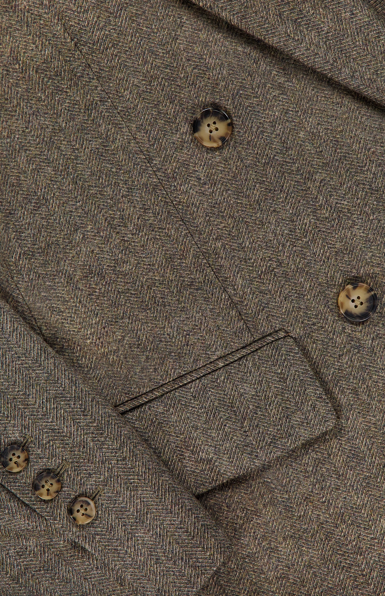Close-up of the Harper Herringbone Blazer by TWP, highlighting the brown herringbone pattern with three brown buttons on the sleeve and three larger buttons on the front, plus a glimpse of a pocket—an elegant detail characteristic of classic herringbone workwear jackets.