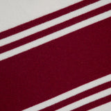 A close-up of TWP’s Hooded Cashmere Stripe Poncho shows its ivory fabric with a diagonal maroon stripe edged by two slim maroon lines, set against an off-white background.