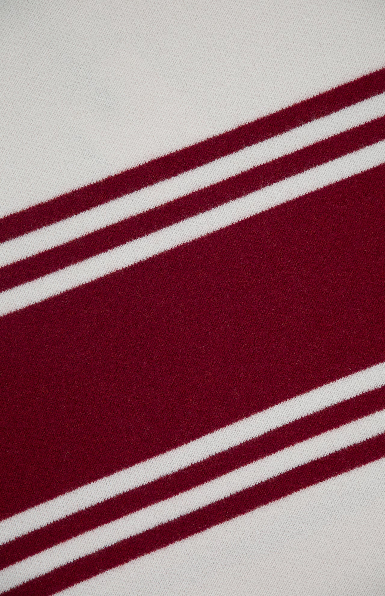 A close-up of TWP’s Hooded Cashmere Stripe Poncho shows its ivory fabric with a diagonal maroon stripe edged by two slim maroon lines, set against an off-white background.