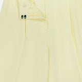 A close-up of the TWP Hudson St Pleated Pant in pale yellow shows the unbuttoned waistband and partially open zipper, revealing striped lining and button details—ideal as relaxed women’s workwear.