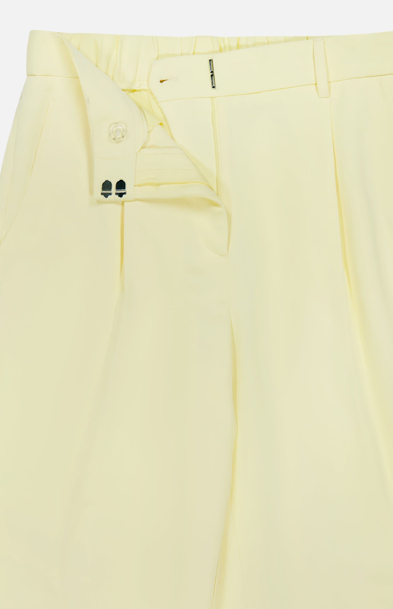 A close-up of the TWP Hudson St Pleated Pant in pale yellow shows the unbuttoned waistband and partially open zipper, revealing striped lining and button details—ideal as relaxed women’s workwear.