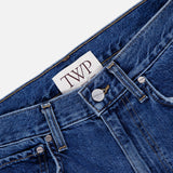 Close-up of the TWP Kent St Jean in Paris, crafted from blue denim and showcasing a silver button, belt loops, a classic five-pocket design with front pockets, and a white inner waistband label featuring bold "TWP" lettering.