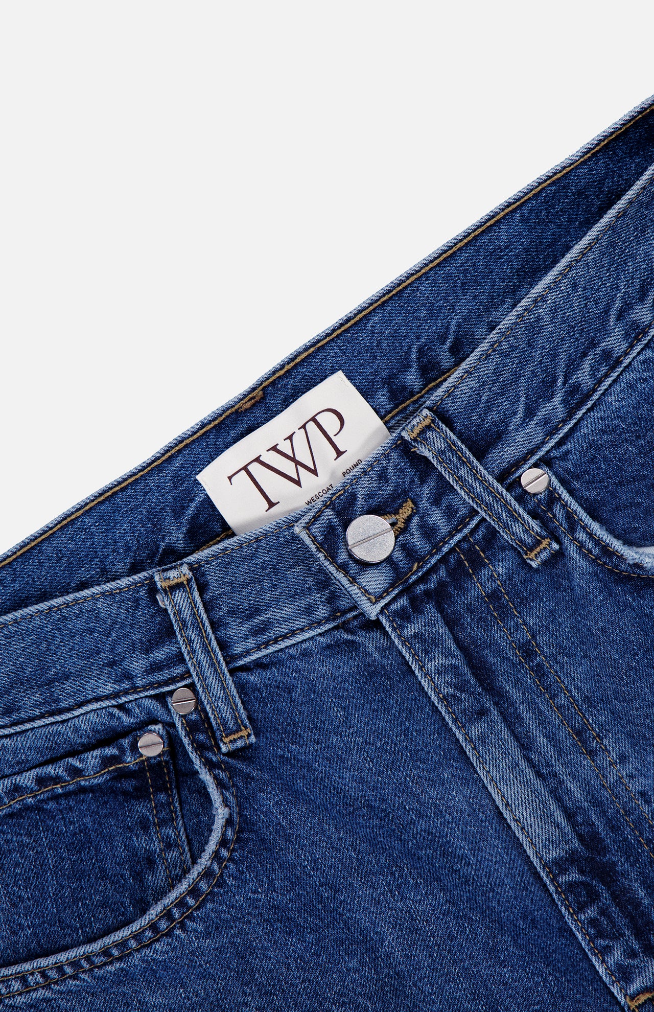 Close-up of the TWP Kent St Jean in Paris, crafted from blue denim and showcasing a silver button, belt loops, a classic five-pocket design with front pockets, and a white inner waistband label featuring bold "TWP" lettering.