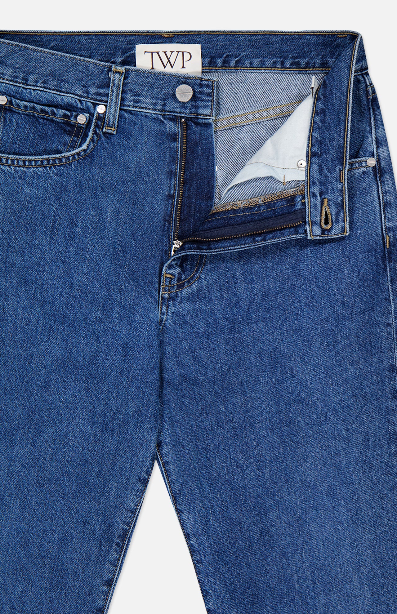 The Kent St Jean in Paris by TWP is a pair of blue jeans with a classic 5-pocket design and straight leg silhouette, featuring a partially unzipped fly that reveals the inner fabric and a waistband label marked with the TWP brand.