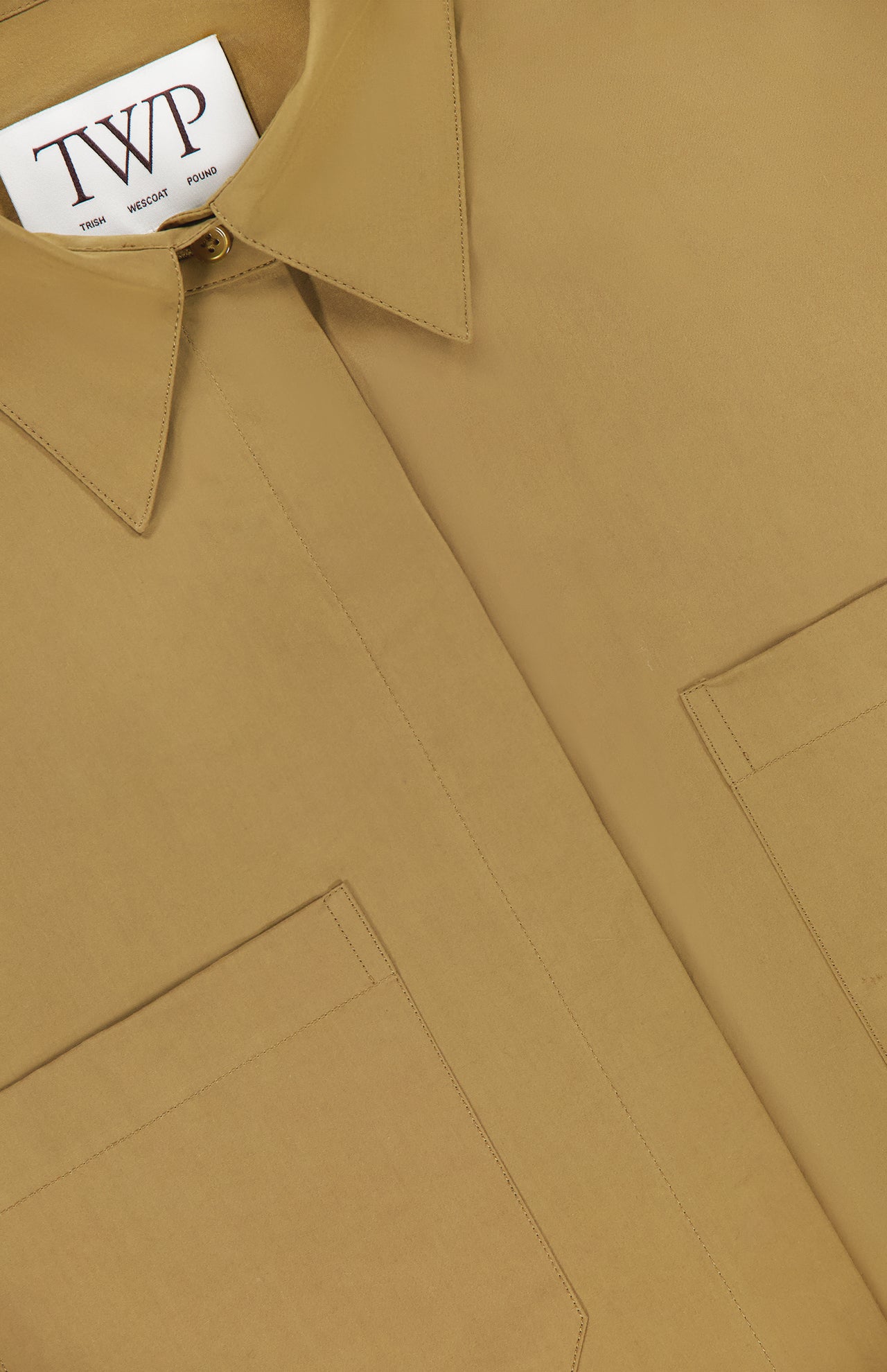 Close-up of the My Newest Mistake Top by TWP—a tan, relaxed fit button-up with two front pockets and a pointed collar. Features a white label with the TWP brand; perfect as a versatile workwear staple for women.