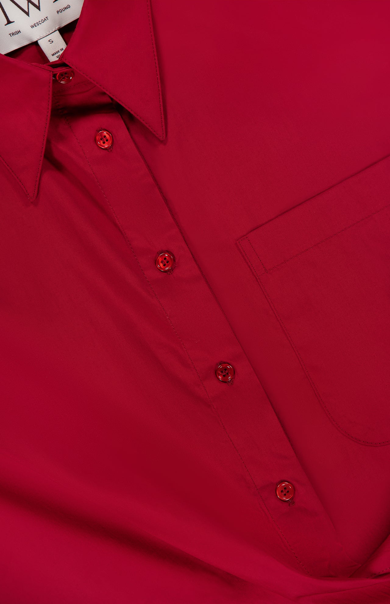 Close-up of the New Earl Wrap Front Dress by TWP: bright red with a breast pocket, red buttons, and pointed collar—ideal women’s workwear. A partially visible clothing label appears inside the collar.