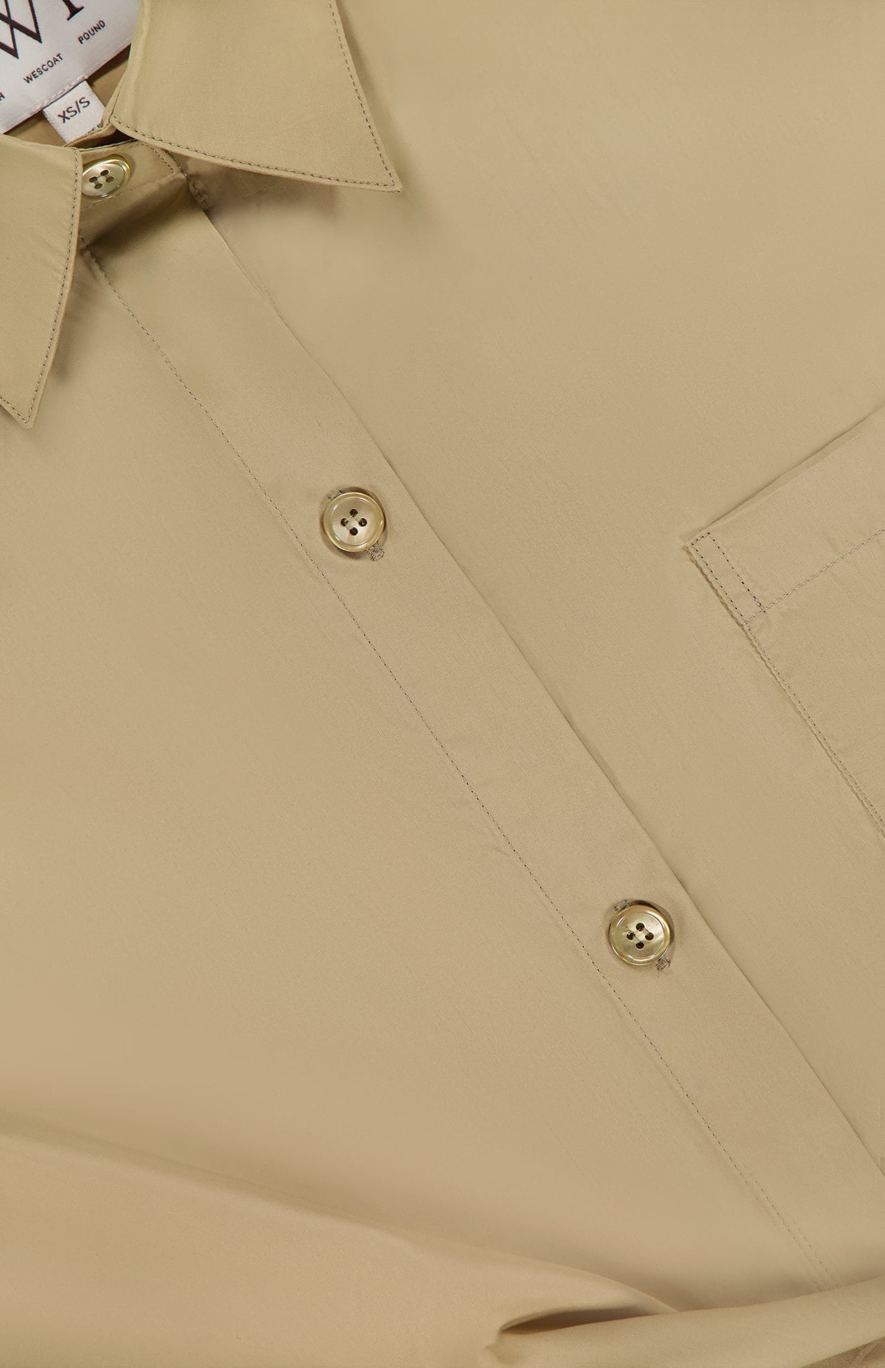 Close-up of the TWP New Earl Asymmetric Button Front Shirt in beige Italian cotton, featuring two buttons, part of a collar, a chest pocket, and a partially visible clothing tag in the upper left corner.