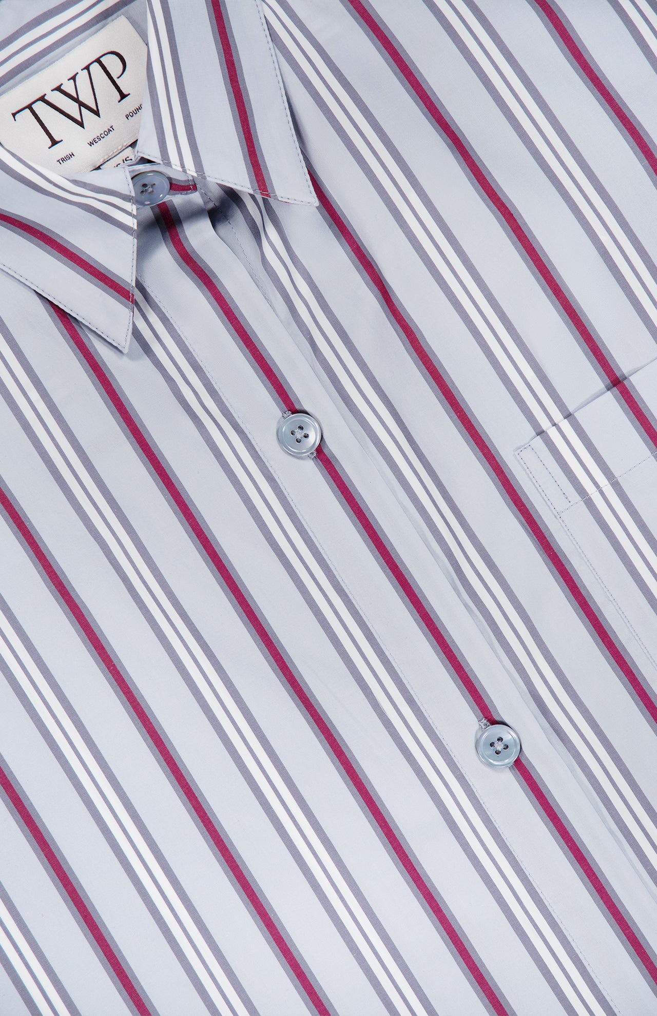 Close-up of the Next Ex Striped Blouse by TWP—a light blue button-up shirt featuring vertical stripes in white, gray, and red for a modern update to women’s workwear. The blouse has a folded collar and a pocket on the right side, combining style with practicality. A label reading TWP is visible inside the collar.