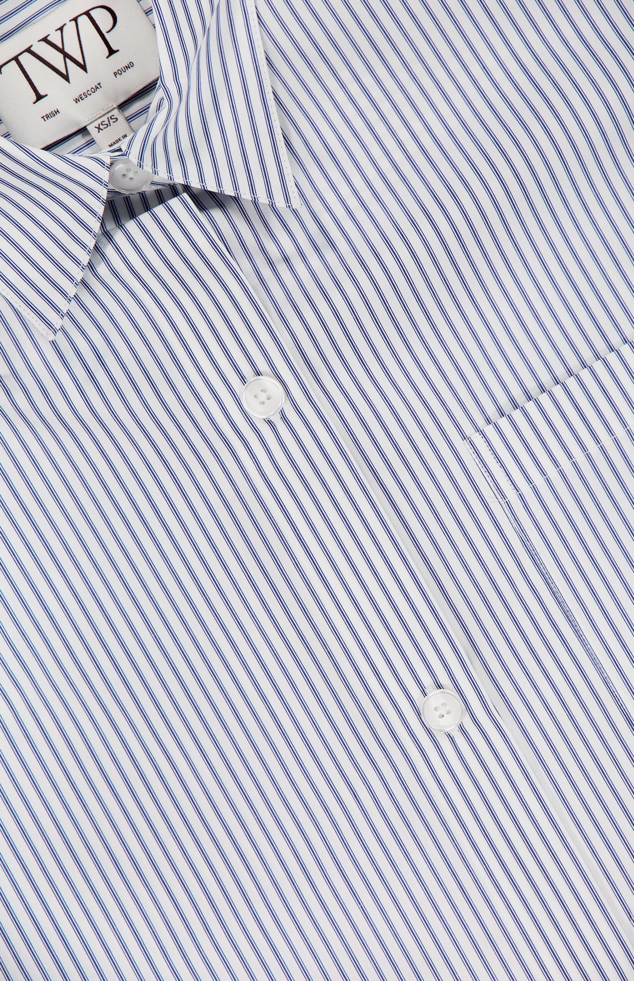 Close-up of the TWP Next Ex Striped Cropped Blouse featuring thin blue vertical stripes, white buttons, a pocket, and a collar—a versatile women’s workwear piece displaying the TWP label.