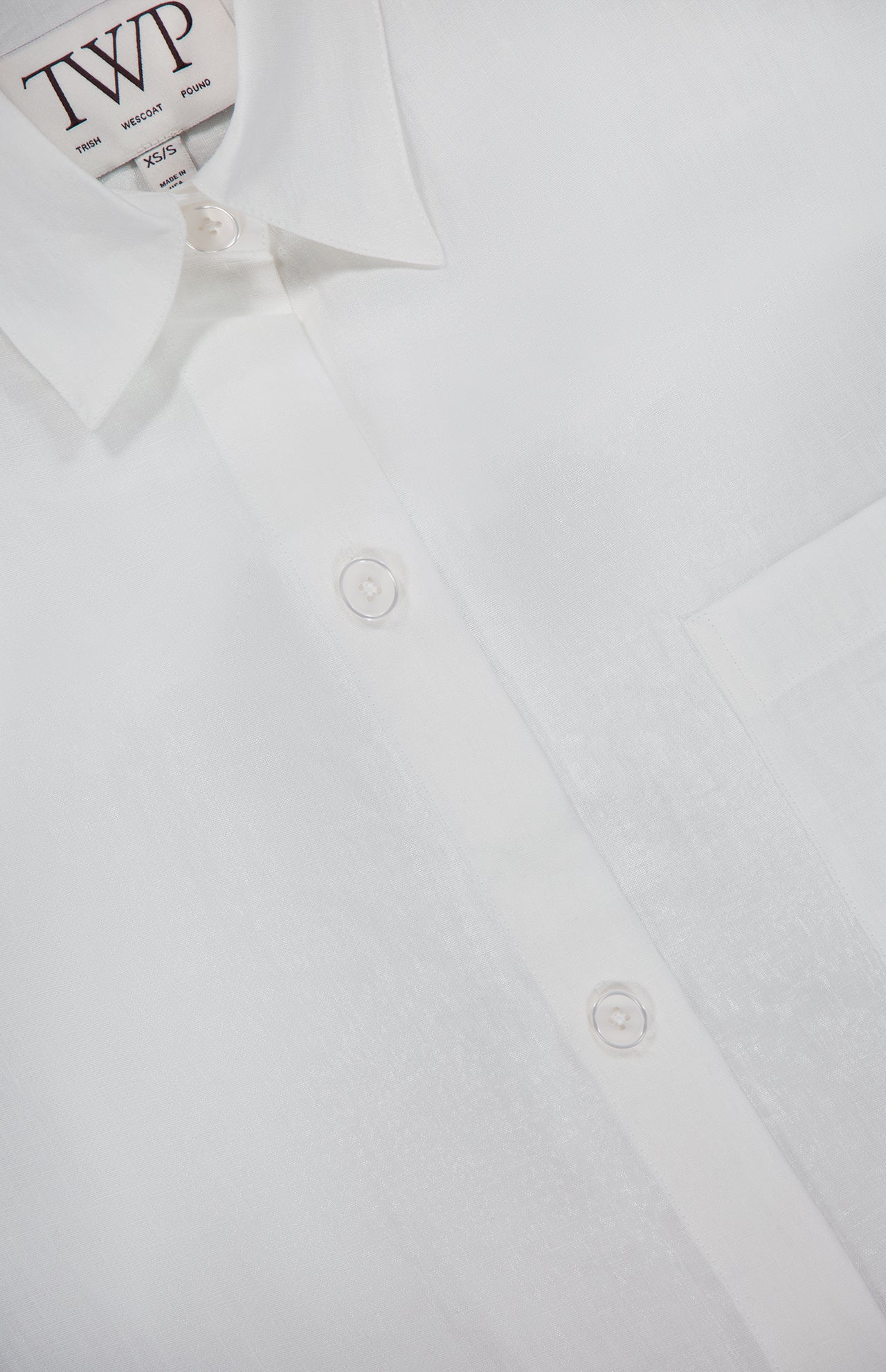 Close-up of the Next Ex Shirt in Linen by TWP—a crisp white, cropped boxy shirt featuring a collar, three white buttons, front pocket, and a modern twist on classic women’s workwear style.