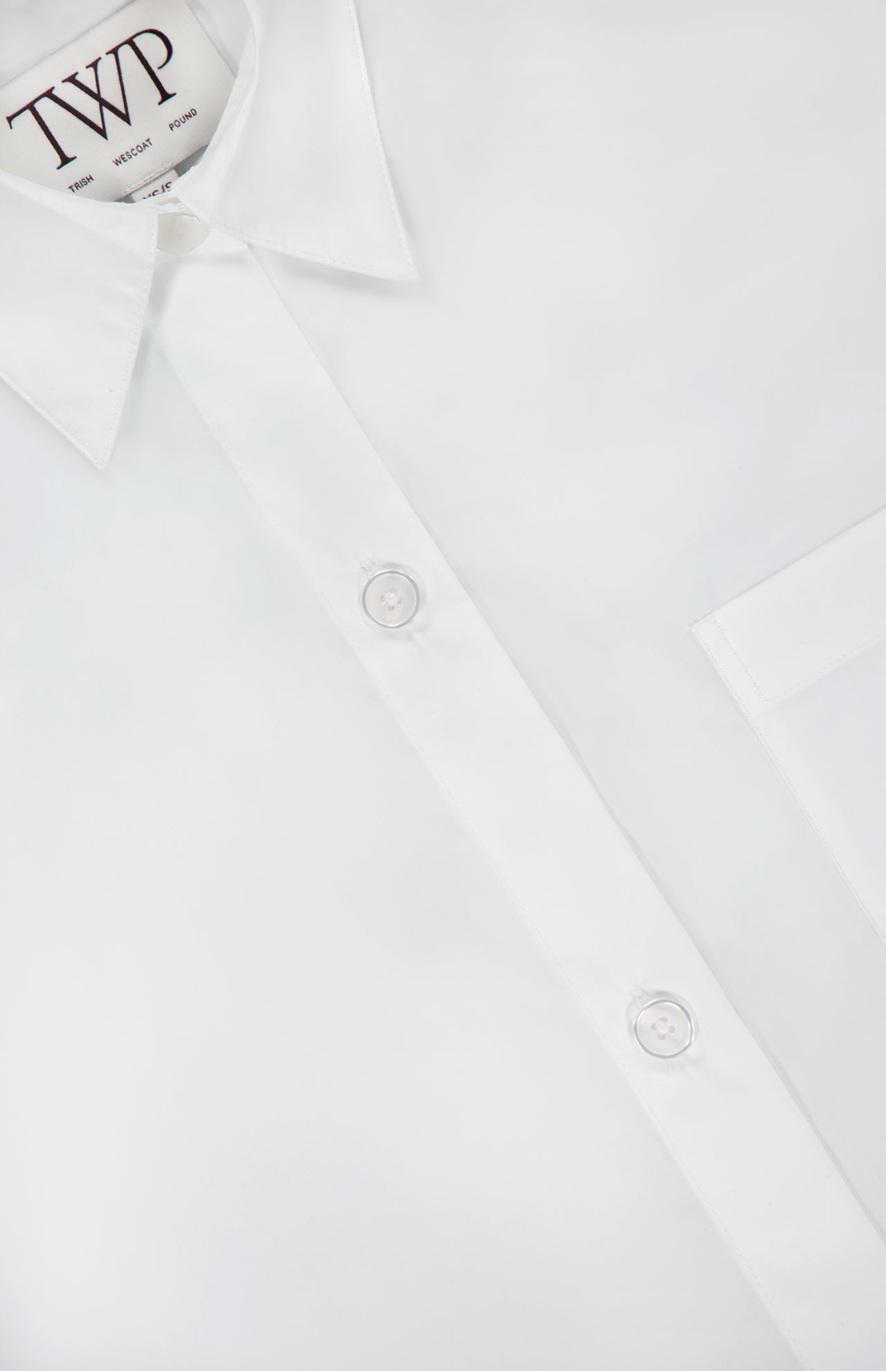 Close-up of the Next Ex Cropped Shirt by TWP—a crisp white, boxy-fit shirt with a pointed collar, chest pocket, and two white buttons. The label reads TWP in black, offering modern elegance for women’s workwear.