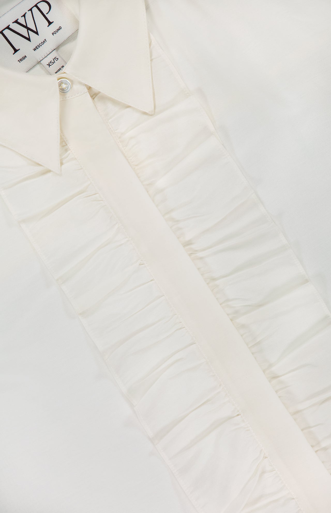 Close-up of the TWP Next Ex Ruffle Blouse in ivory white, featuring ruffled detailing along the button placket. Made from smooth, lightweight fabric, this medium-sized blouse blends comfort and sophistication—perfect for women's workwear.