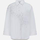 The TWP Next Morning Crystal Shirt is a white oversized Italian cotton shirt with long sleeves and a chest pocket, adorned with a cluster of small silver studs that form embellished crystal detailing on the left shoulder and upper chest—offering both comfort and a modern, elevated edge.