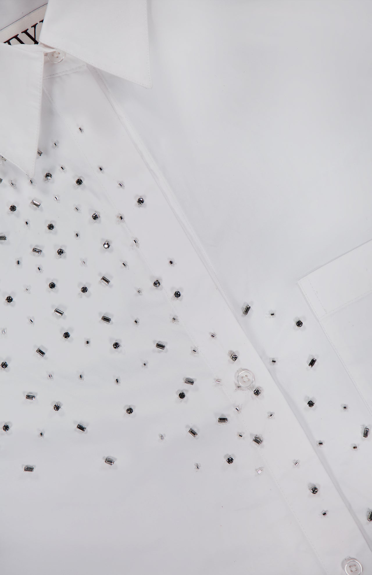 Close-up of the TWP Next Morning Crystal Shirt, an oversized white shirt expertly crafted from fine Italian cotton. The design is adorned with small, reflective silver crystal embellishments concentrated mostly on one side, and features classic buttons, a visible collar, and subtle sparkling details for an elevated finish.