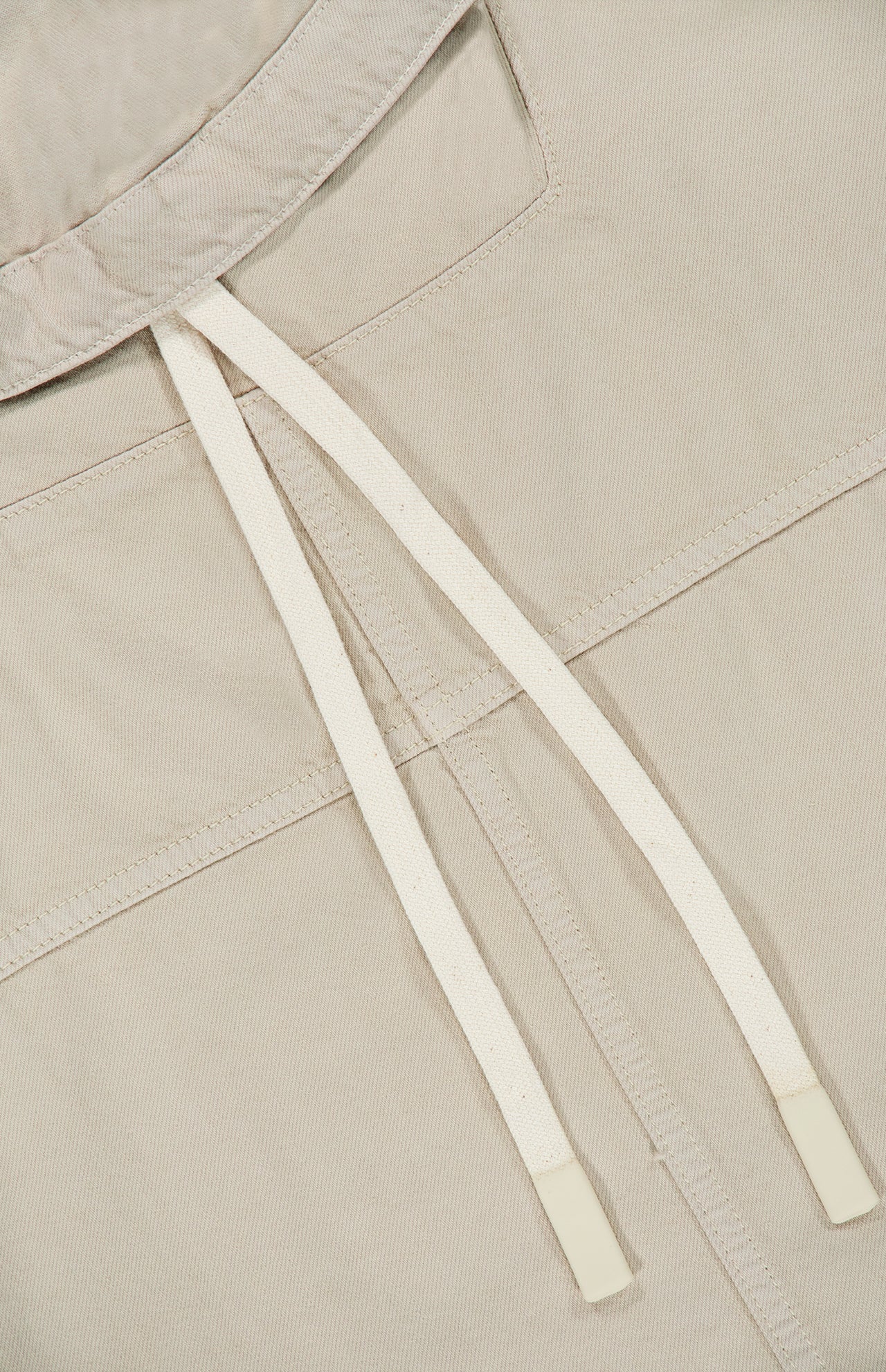 Close-up of the Pull Me Over Jacket by TWP in beige, featuring a V-shaped collar and two white, plastic-tipped drawstrings. The textured fabric and functional details elevate this women’s workwear staple with modern style.