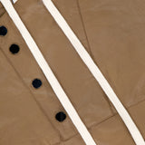 Close-up of the TWP Water-repellent Rain Check Jacket in brown leather-like cotton with white drawstrings, black snap buttons, visible stitching, and a partially visible white tag with black text in the background.