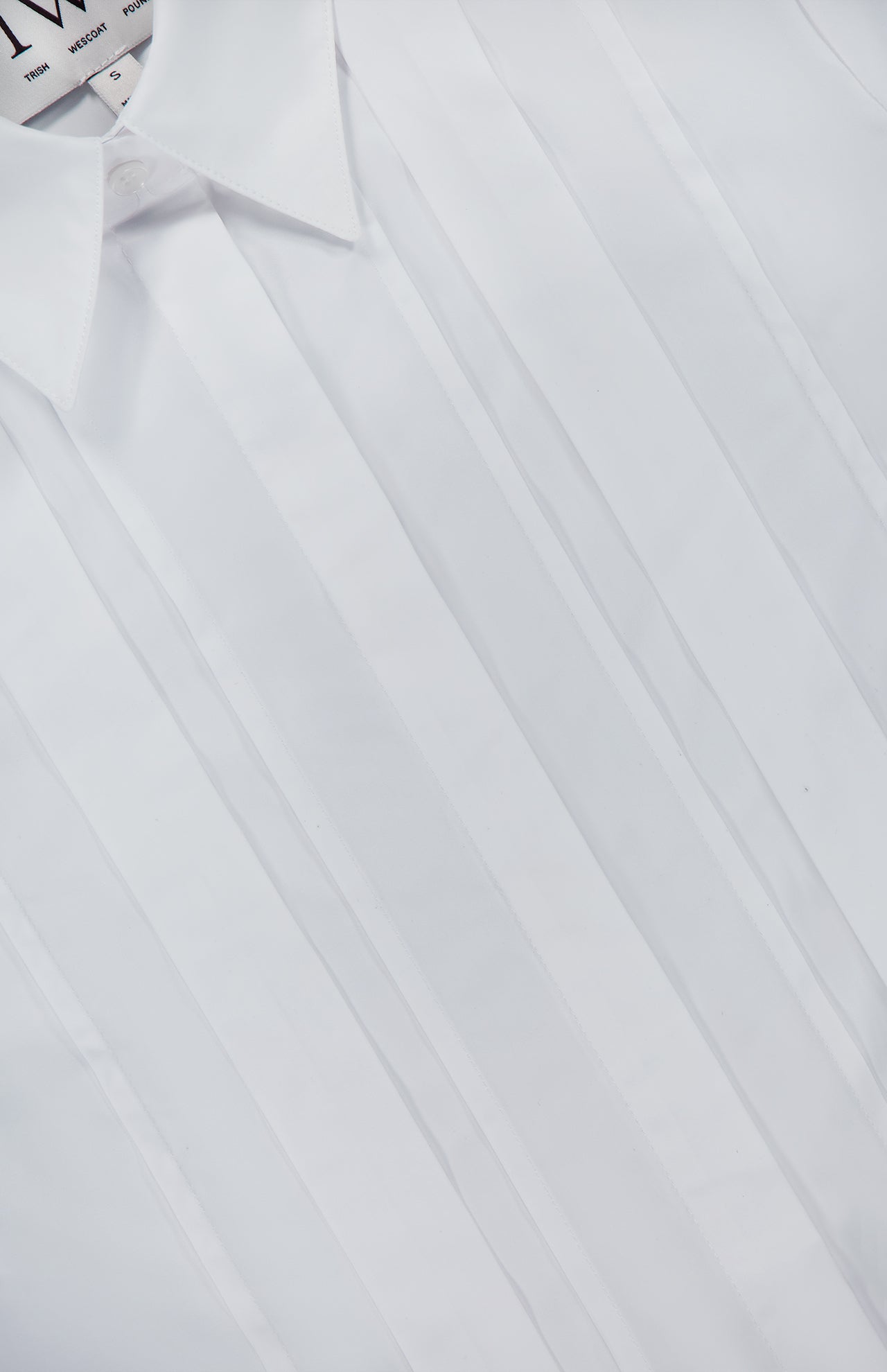 Close-up of the TWP Short Lived Placket with Cherry Pleats Shirt, a cropped white dress shirt featuring vertical pleats and a pointed collar. Neatly pressed and laid flat with the top button undone, this piece showcases a modern update on classic women’s workwear. Part of the inside label is visible, emphasizing its refined design.