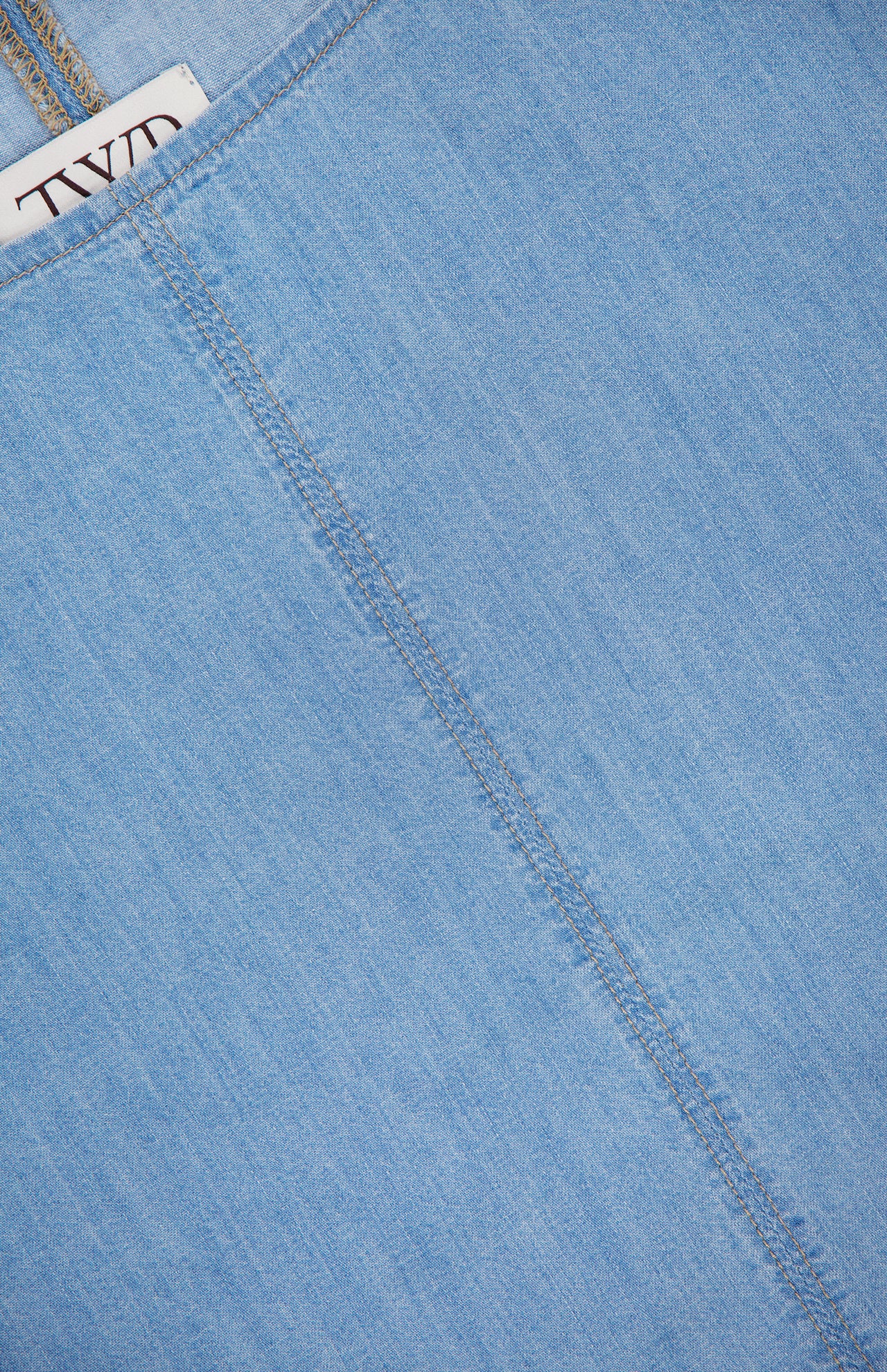 Close-up of the Simple T Top by TWP in lightweight denim, featuring a diagonal seam and a partially visible white label with black text—a modern take on women’s workwear style.