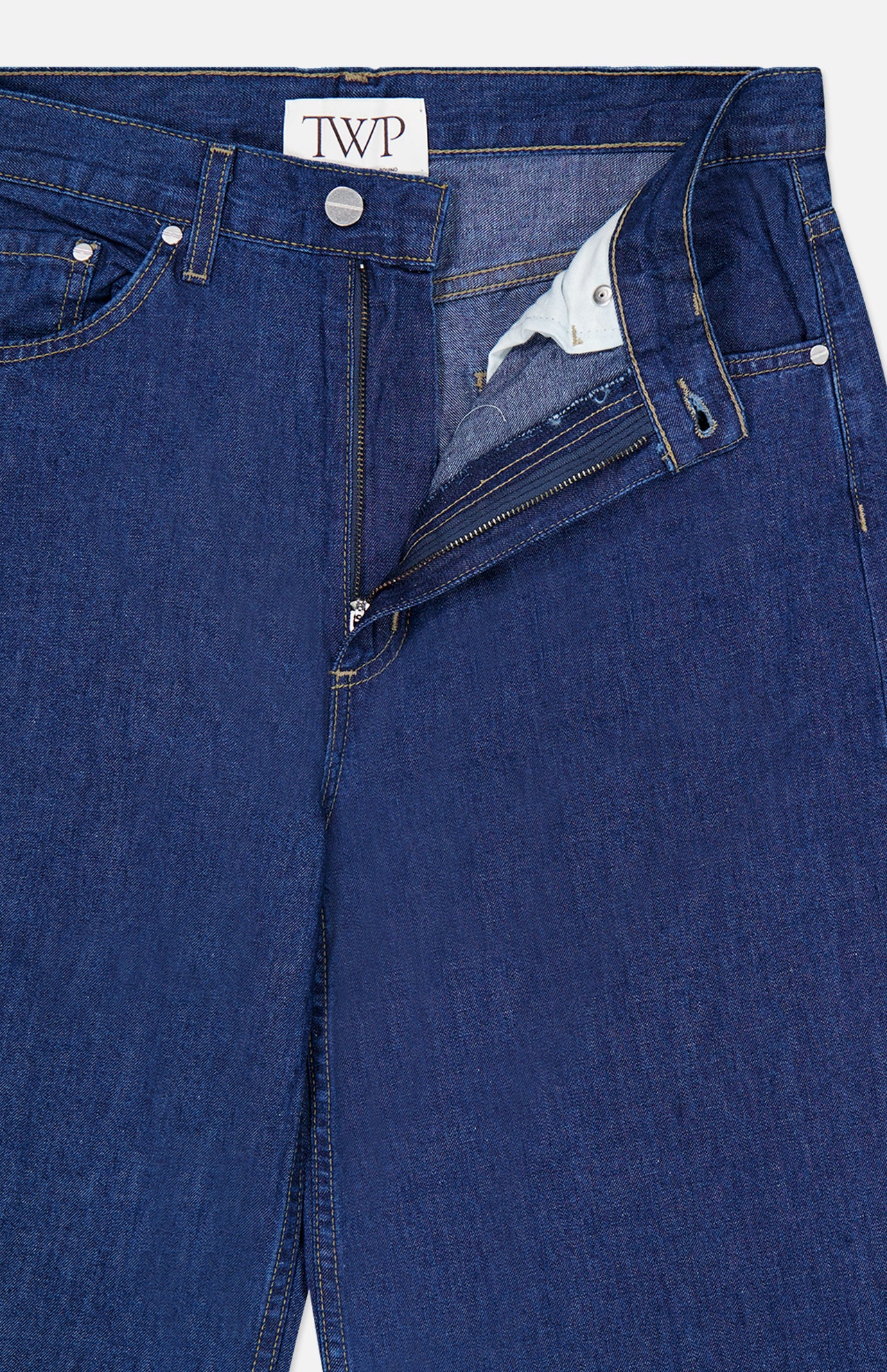 Close-up of the Song Sung Blue Jean in Dark Stone by TWP, featuring relaxed blue denim with a partially open zipper and button fly that reveals the inner pocket lining and a TWP label on the waistband.