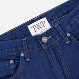 Close-up of the Song Sung Blue Jean in Dark Stone by TWP, crafted from a blue denim cotton linen blend with visible stitching, metal rivets, belt loops, and a white label on the waistband featuring the bold TWP logo.