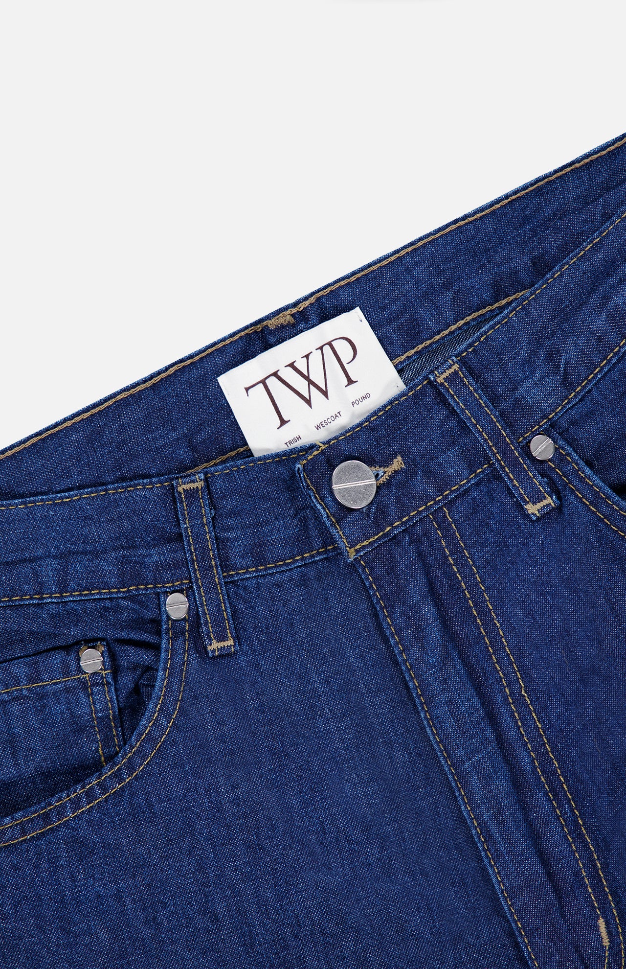 Close-up of the Song Sung Blue Jean in Dark Stone by TWP, crafted from a blue denim cotton linen blend with visible stitching, metal rivets, belt loops, and a white label on the waistband featuring the bold TWP logo.