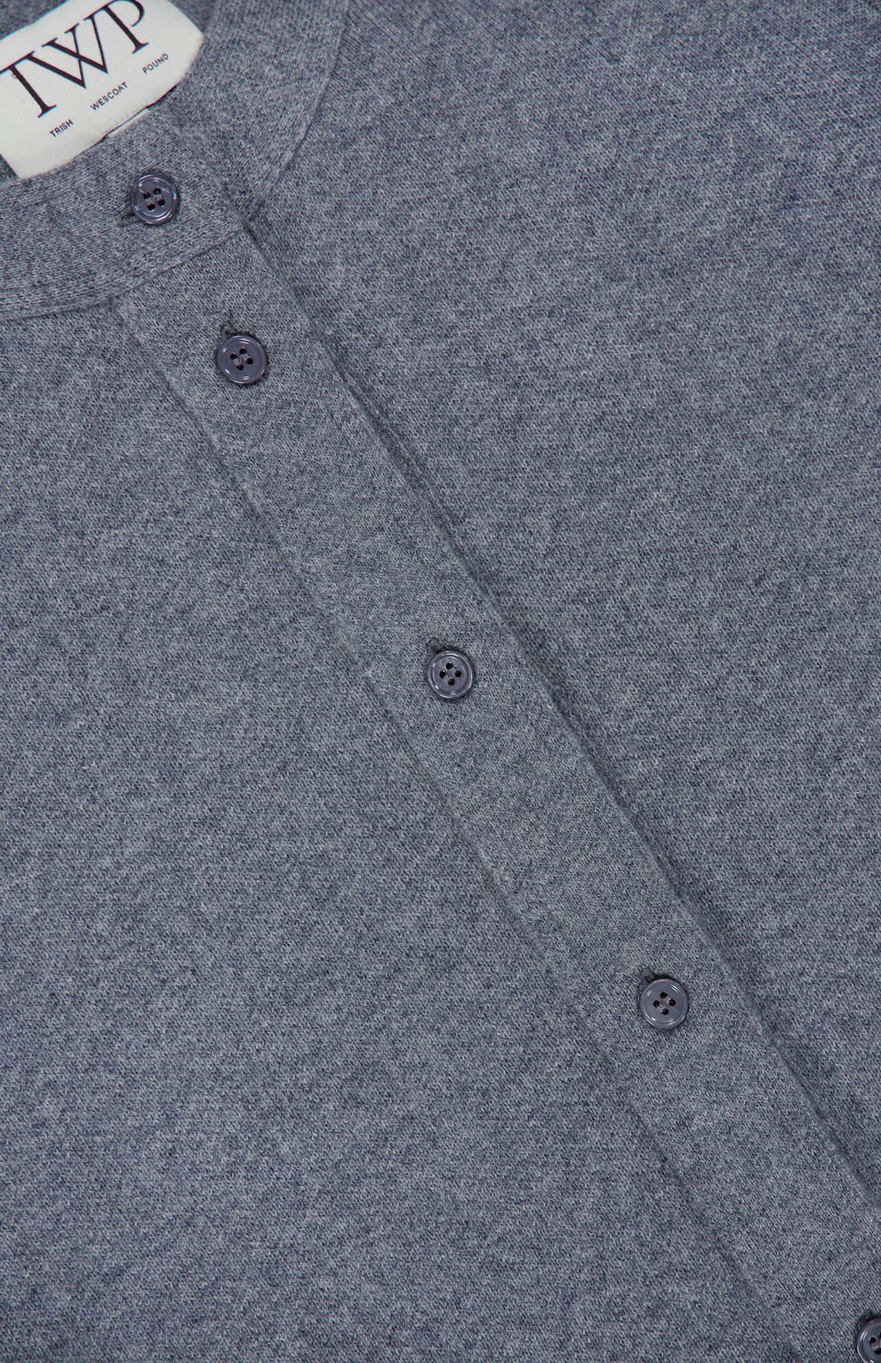 Close-up of a heather gray Thea Fringed Hem Cardigan from TWP, featuring four dark buttons and a visible TWP brand label at the collar. The fabric has a soft, textured appearance reminiscent of Italian cashmere, making it an elegant addition to women’s workwear.
