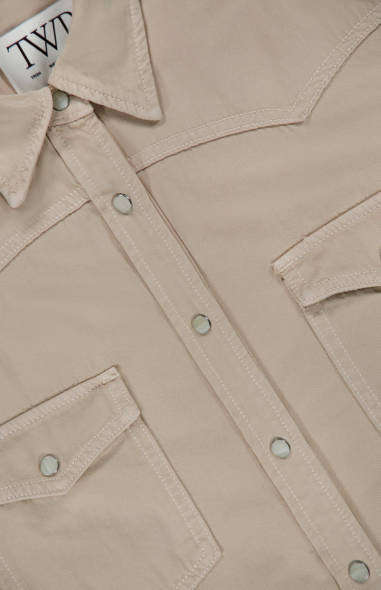 Close-up of the TWP Yellowstone Western Blouse in beige, featuring silver snap buttons, two chest flap pockets, and visible white stitching. A portion of the inner collar label with black text is also visible.