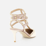 The VALENTINO GARAVANI Rockstud Ankle Strap Heel T.65 in beige patent leather features three ankle straps, gold rockstud details, and a pointed toe, shown against a white background.