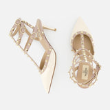The VALENTINO GARAVANI Rockstud Ankle Strap Heel T.65 features nude patent leather, caged pointed toes, iconic rockstuds, ankle straps with buckle closures, and is shown on a white background.